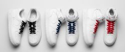 Flat-lay studio shot of three pairs of classic white Nike Air Force 1 sneakers—Low, Mid, and High-top—each featuring different colored laces (black, navy, and red) on a light grey background.